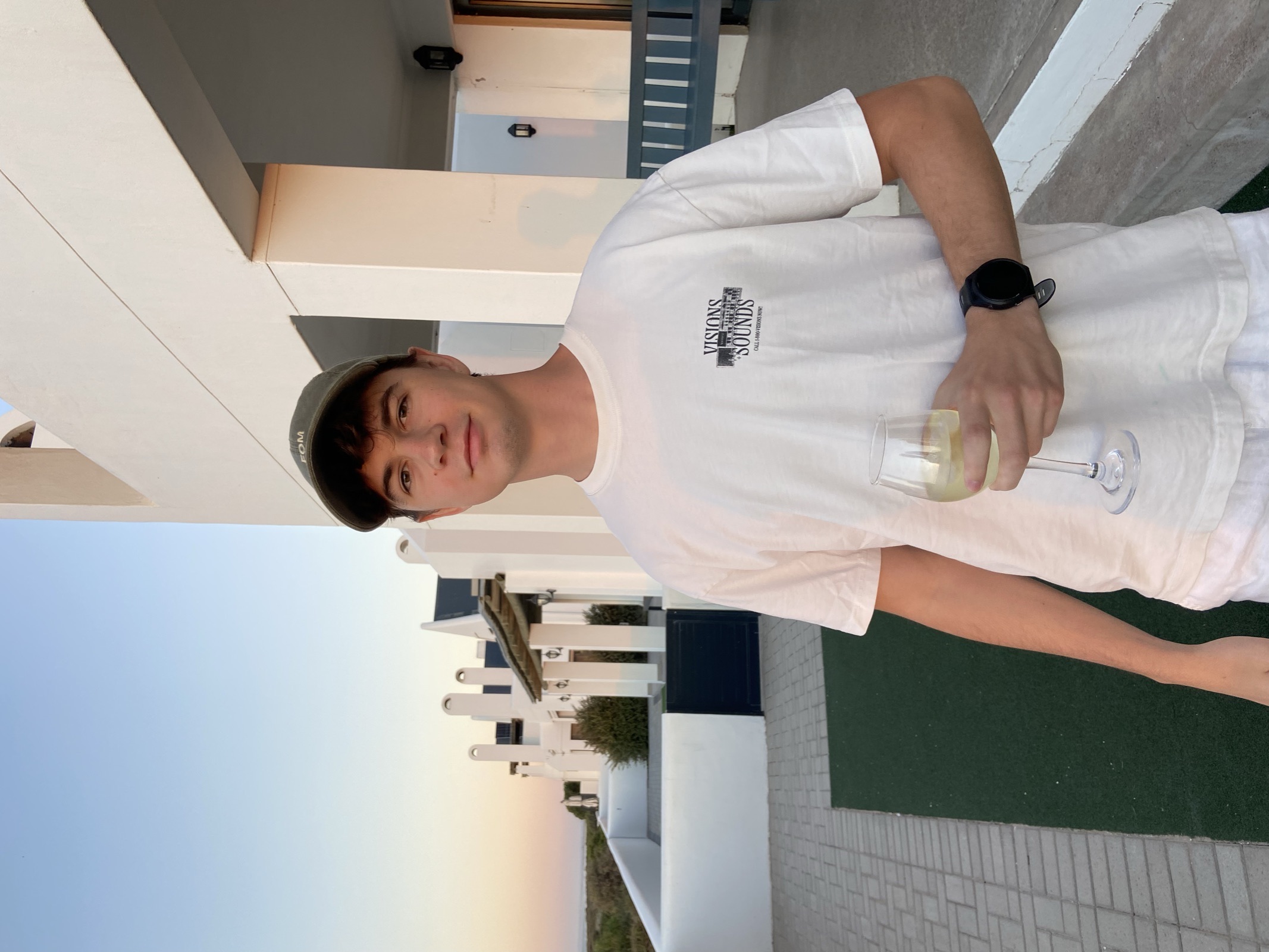 Brendon Lock in a cap and white t-shirt, holding a glass of wine outside a white building at dusk